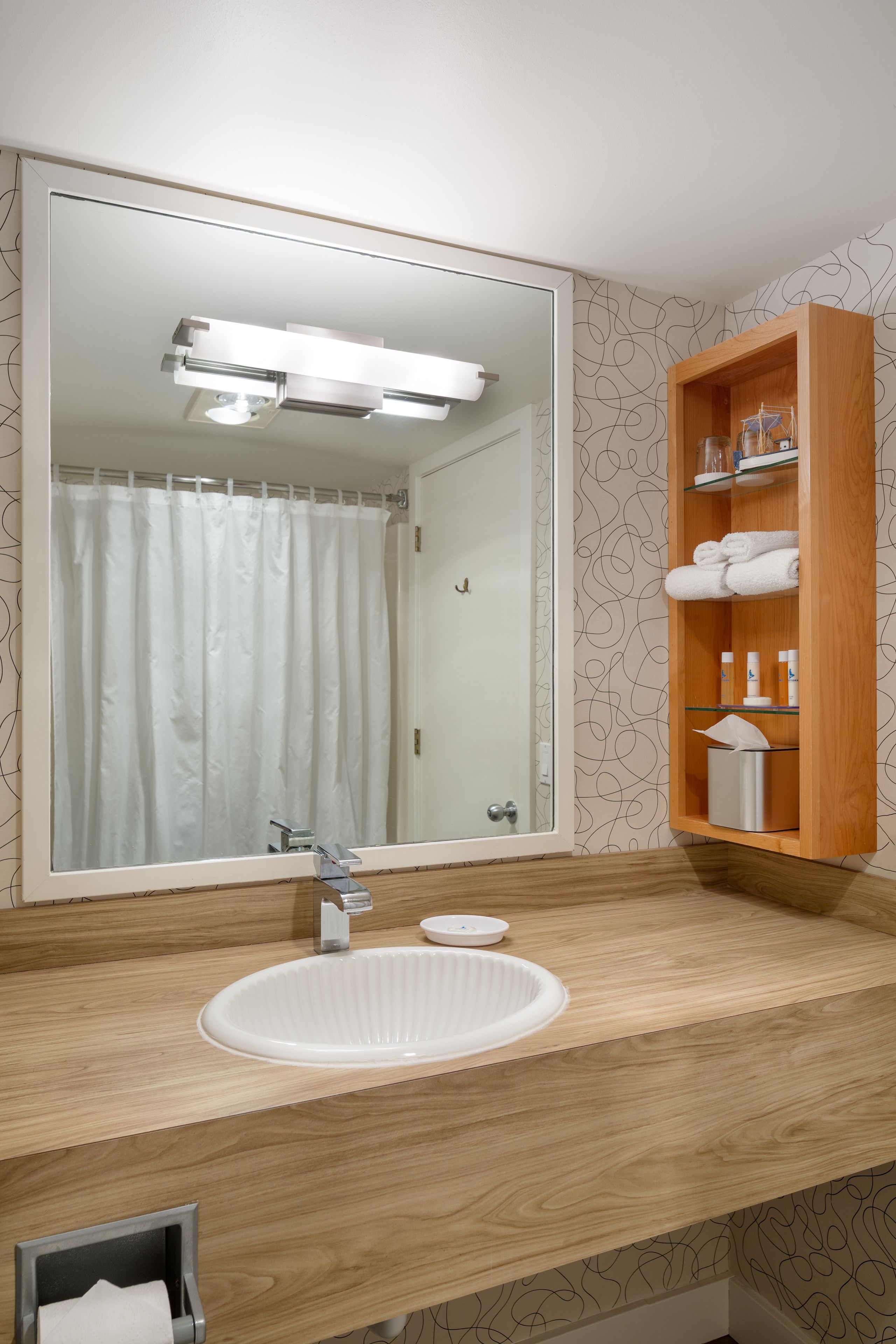 Une salle de bain avec un grand miroir, un meuble-lavabo en bois, un lavabo blanc et une étagère contenant des articles de toilette.