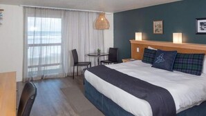 Een hotelkamer met een groot bed, een bureau en een stoel. Er is een raam met gordijnen, een lamp en een schilderij aan de muur.