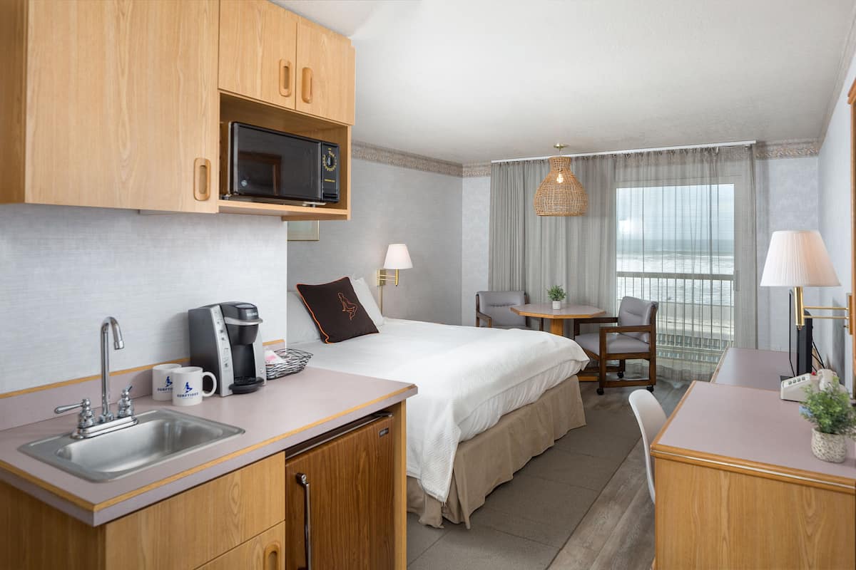 Une chambre d’hôtel avec une kitchenette, un lit recouvert d’une couette blanche, un bureau avec une lampe et une vue sur la mer par la fenêtre.