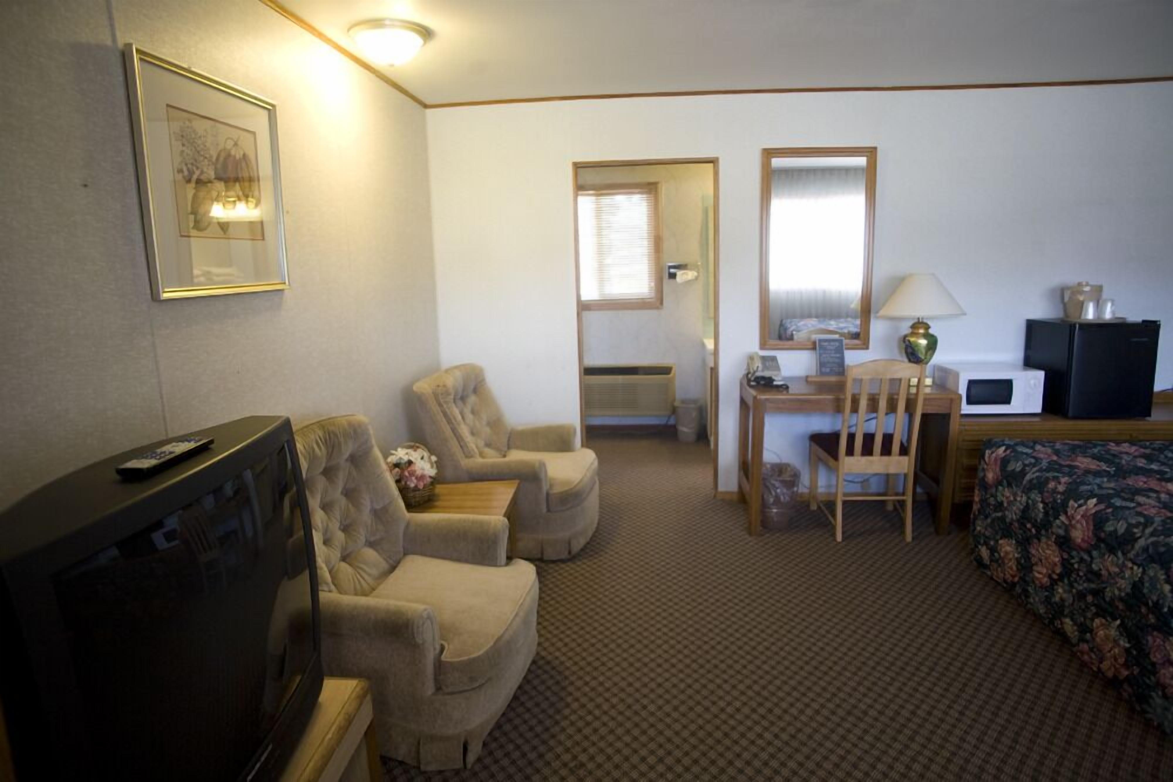 A hotel room with a television, two armchairs, a desk with a chair, a bed with a floral bedspread, and a mirror.