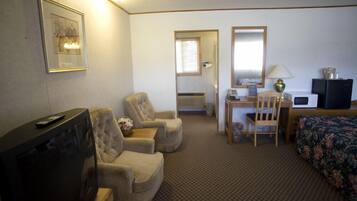 A hotel room with a television, two armchairs, a desk with a chair, a bed with a floral bedspread, and a mirror.