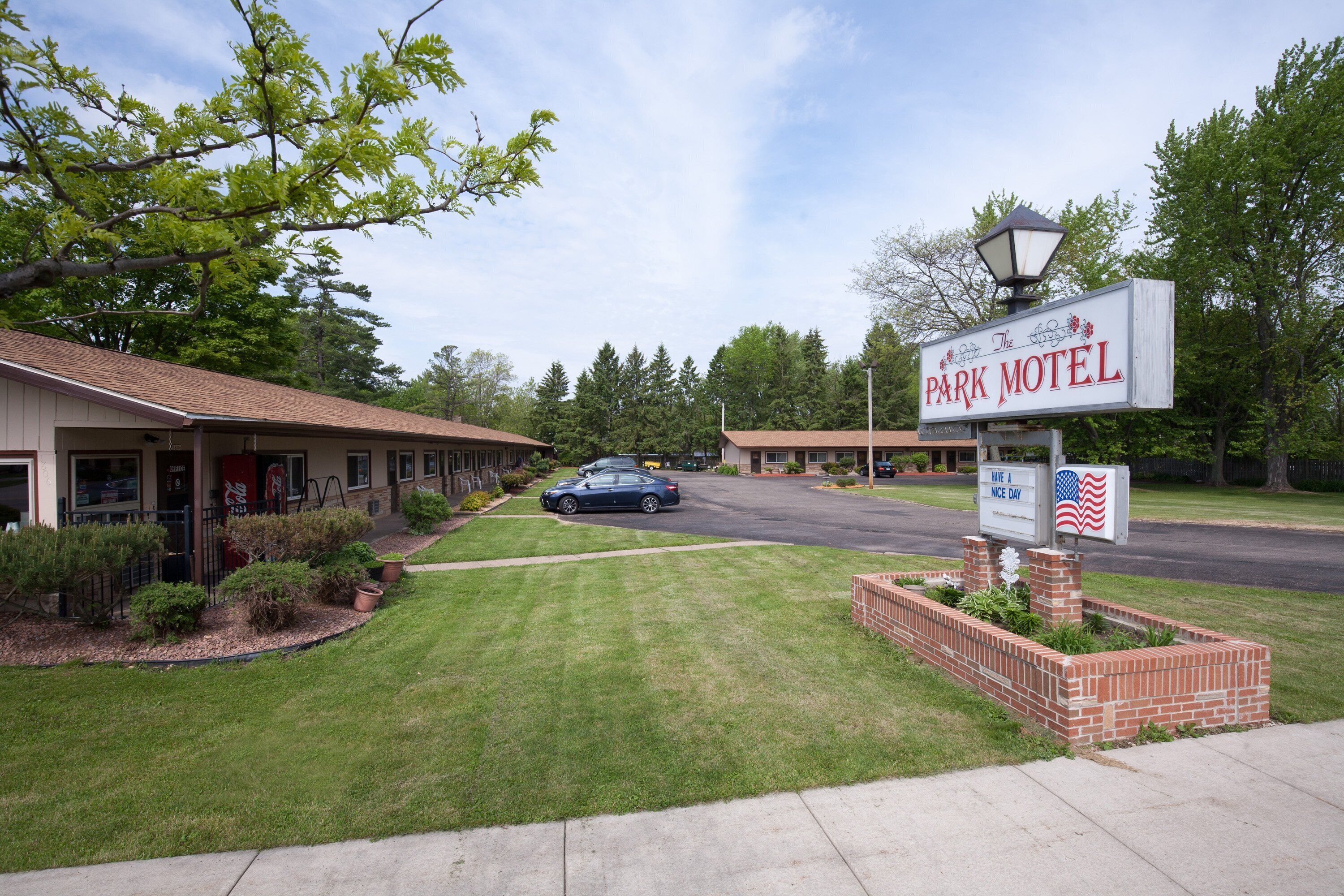 A motel with a sign reading "The Park Motel" and an American flag. There are two buildings, a parking lot, and a tree in the foreground.
