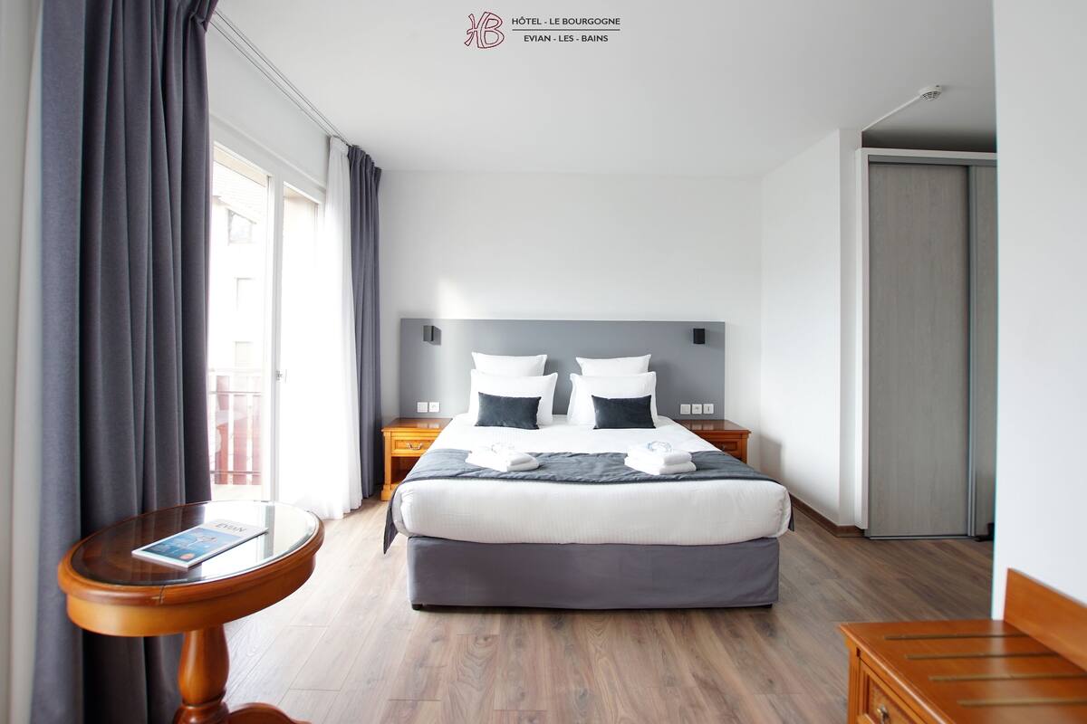 A hotel room with a bed, bedside tables, a small table with a book, and a view of the outside.