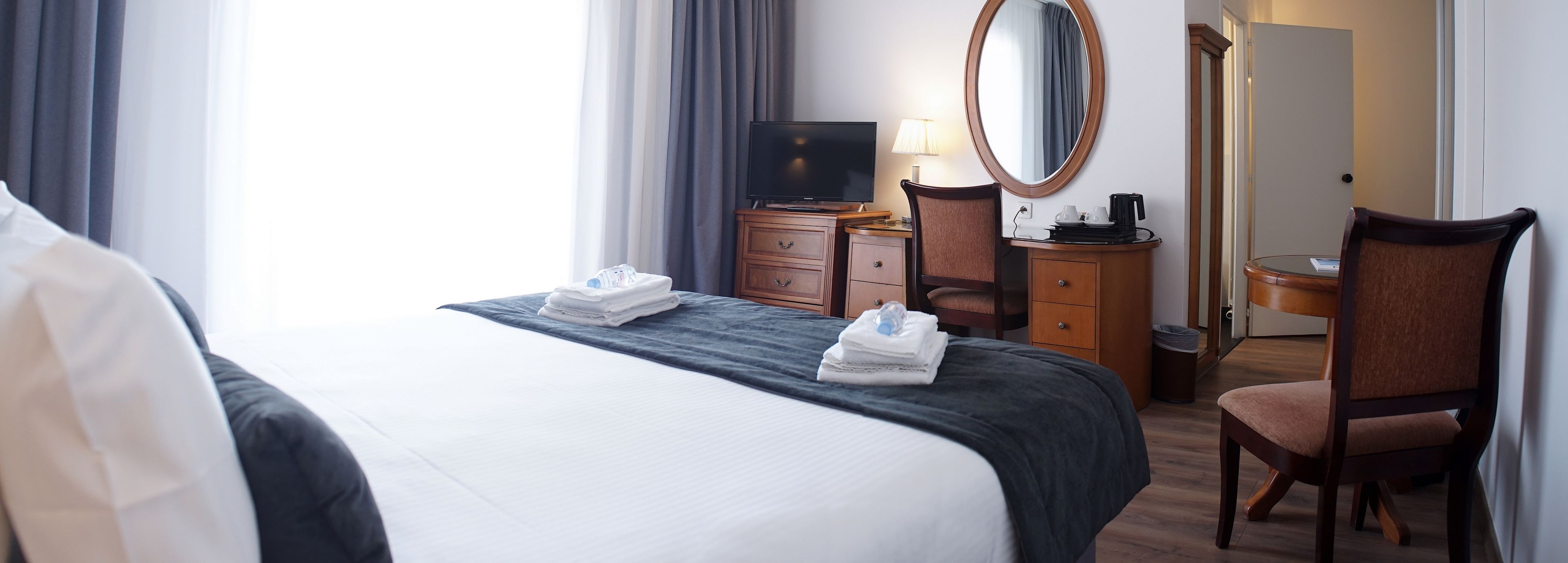 Et hotelværelse med en seng, et skrivebord med computer, en stol, et spejl og et lille bord med lampe.