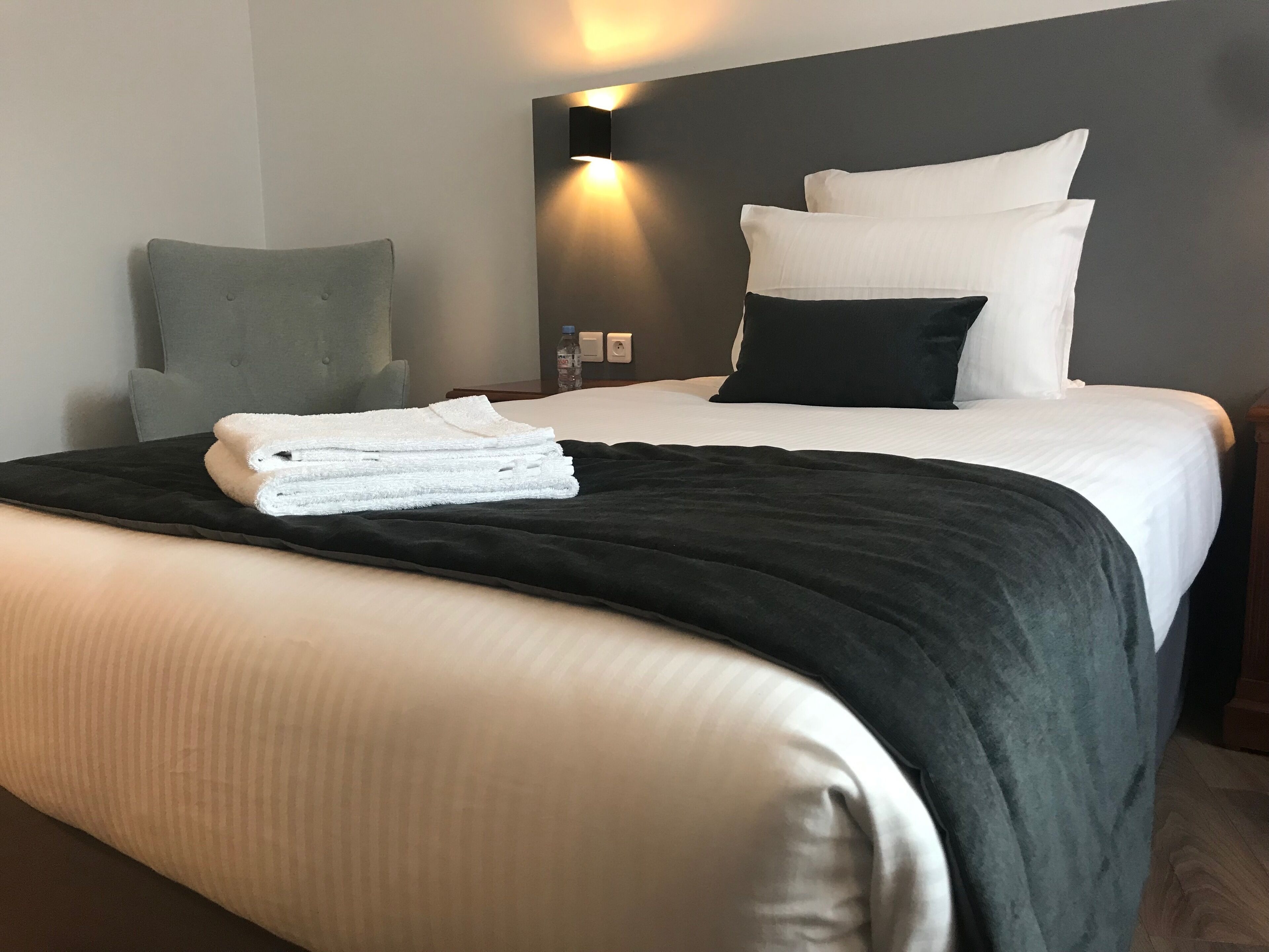 A neatly made bed with white and black bedding, a grey armchair, and a wall-mounted light fixture.