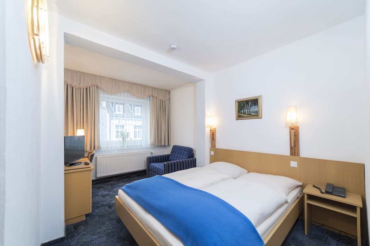 A hotel room with a bed, a desk with a computer, a chair, a window with curtains, and a painting on the wall.