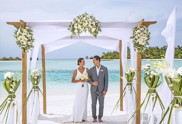 A couple standing under a white floral arch on a beach with clear turquoise water and overwater bungalows in the background. - Naladhu Private Island Maldives (Naladhu)