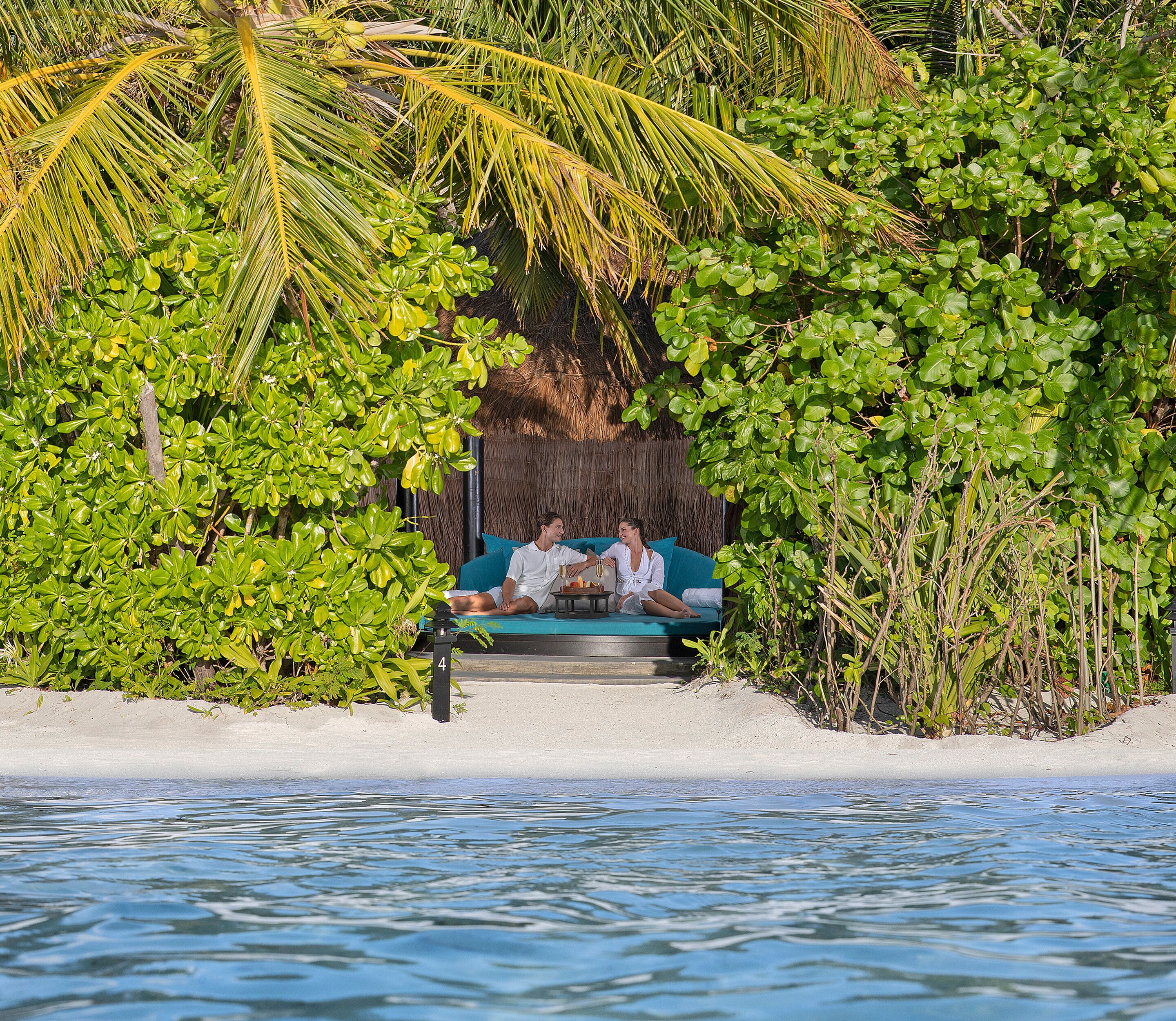 Una pareja descansando en un sofá azul bajo una palmera, rodeada de exuberante vegetación y una playa arenosa.