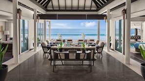 A modern outdoor dining area with a view of the ocean, featuring a long table with chairs, potted plants, and a glass partition.