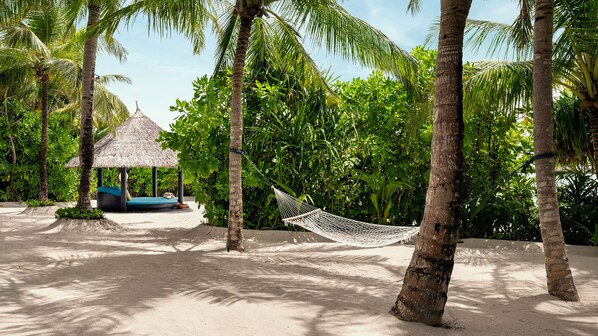 A hammock between palm trees on a sandy beach with a thatched-roof hut and a blue lounge chair in the background.