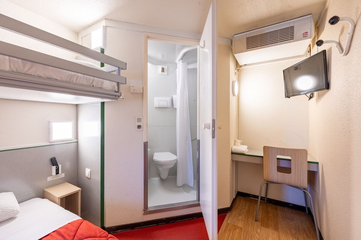 Een kleine hotelkamer met een stapelbed, een douchegedeelte, een klein bureau met stoel en een wandgemonteerde televisie.