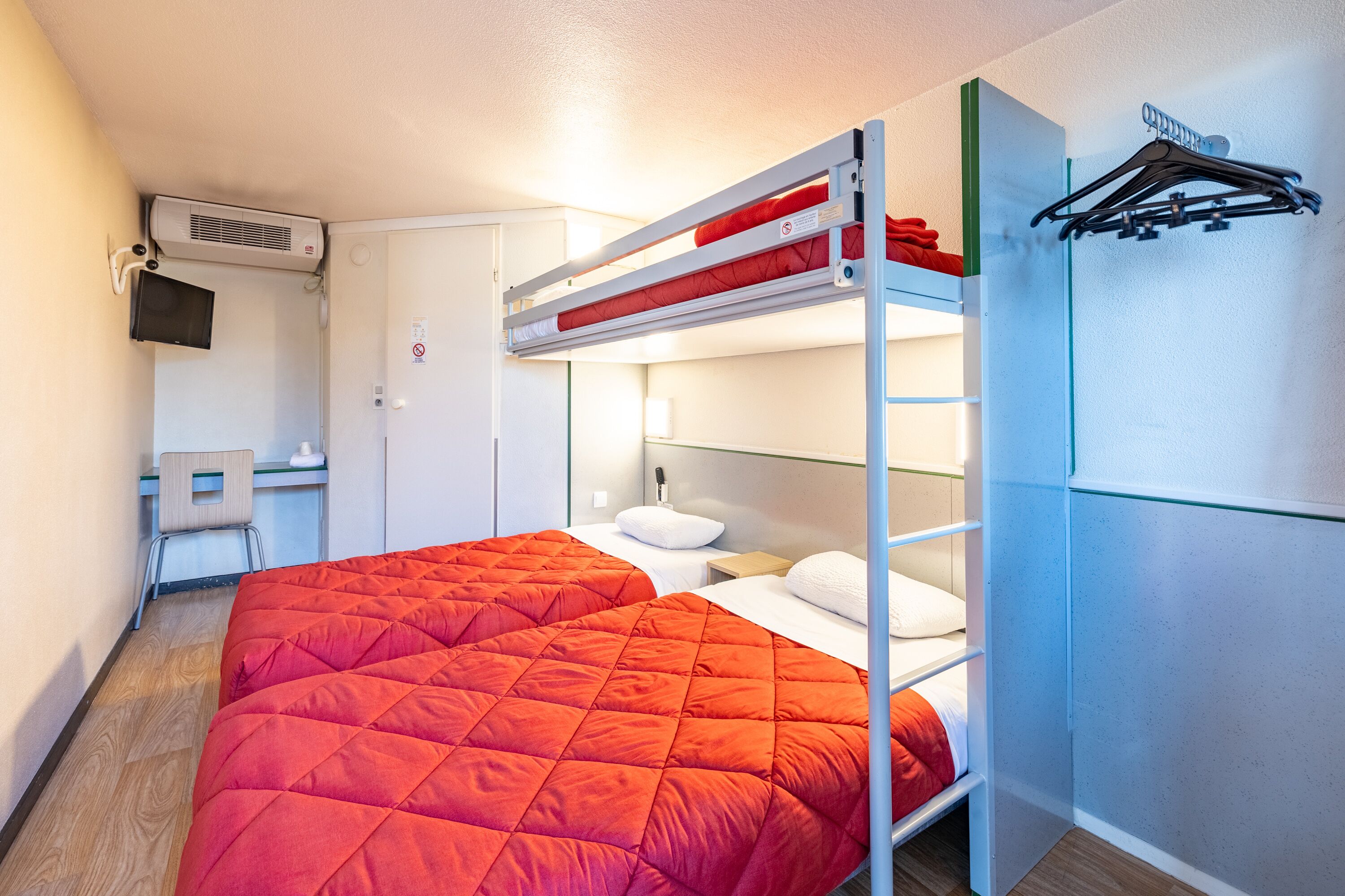 A bunk bed with a red quilt, a desk with a chair, a television, and a wall-mounted heater.