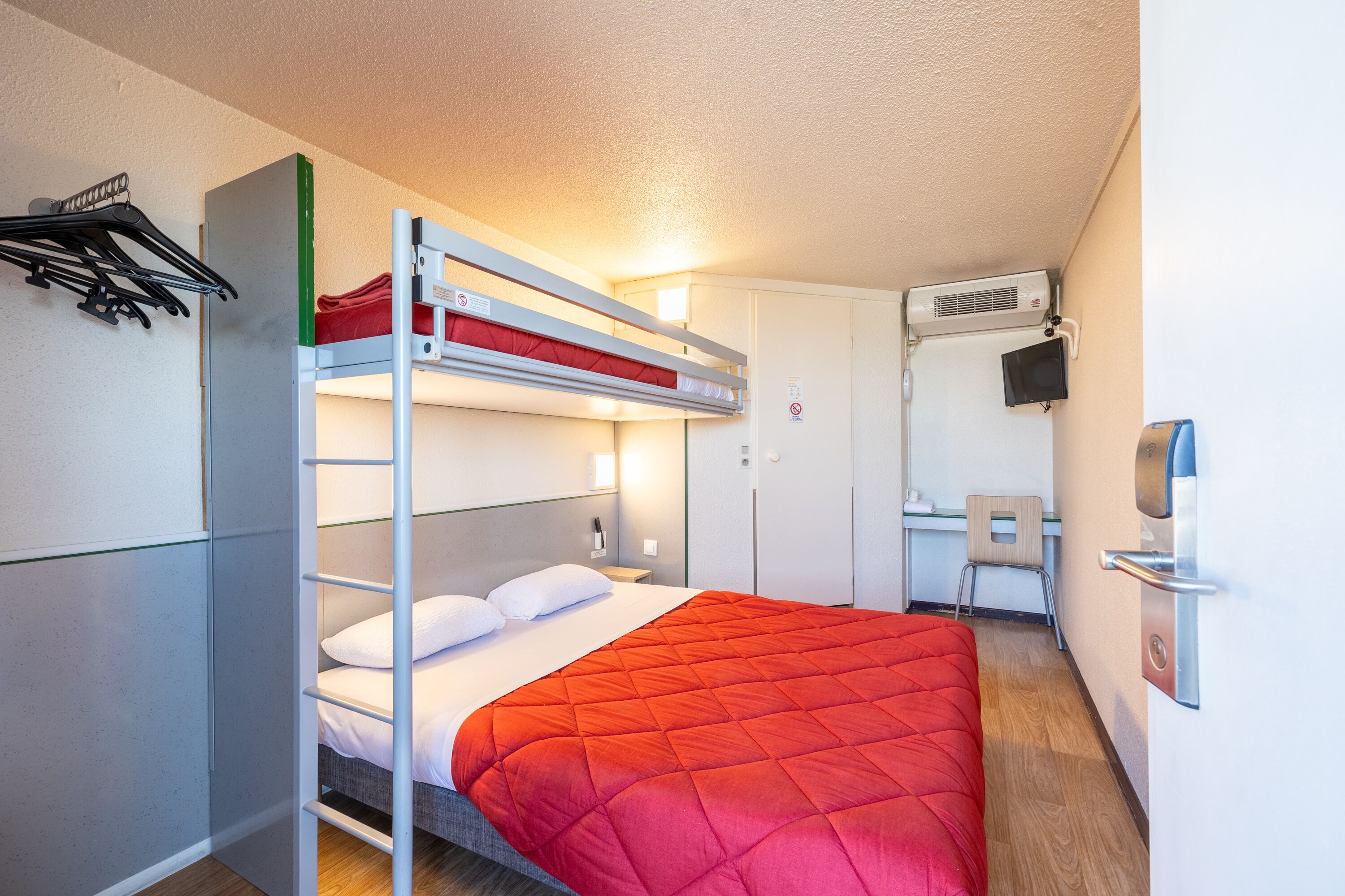 Ein kompaktes Hotelzimmer mit Etagenbett, Schreibtisch mit Stuhl, Fernseher und Kleiderhaken.