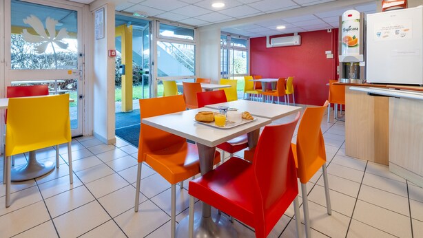 Une cafétéria bien éclairée, avec des chaises orange et jaunes, une table blanche où sont disposés des aliments pour le petit-déjeuner, et un réfrigérateur équipé d’un distributeur d’eau.