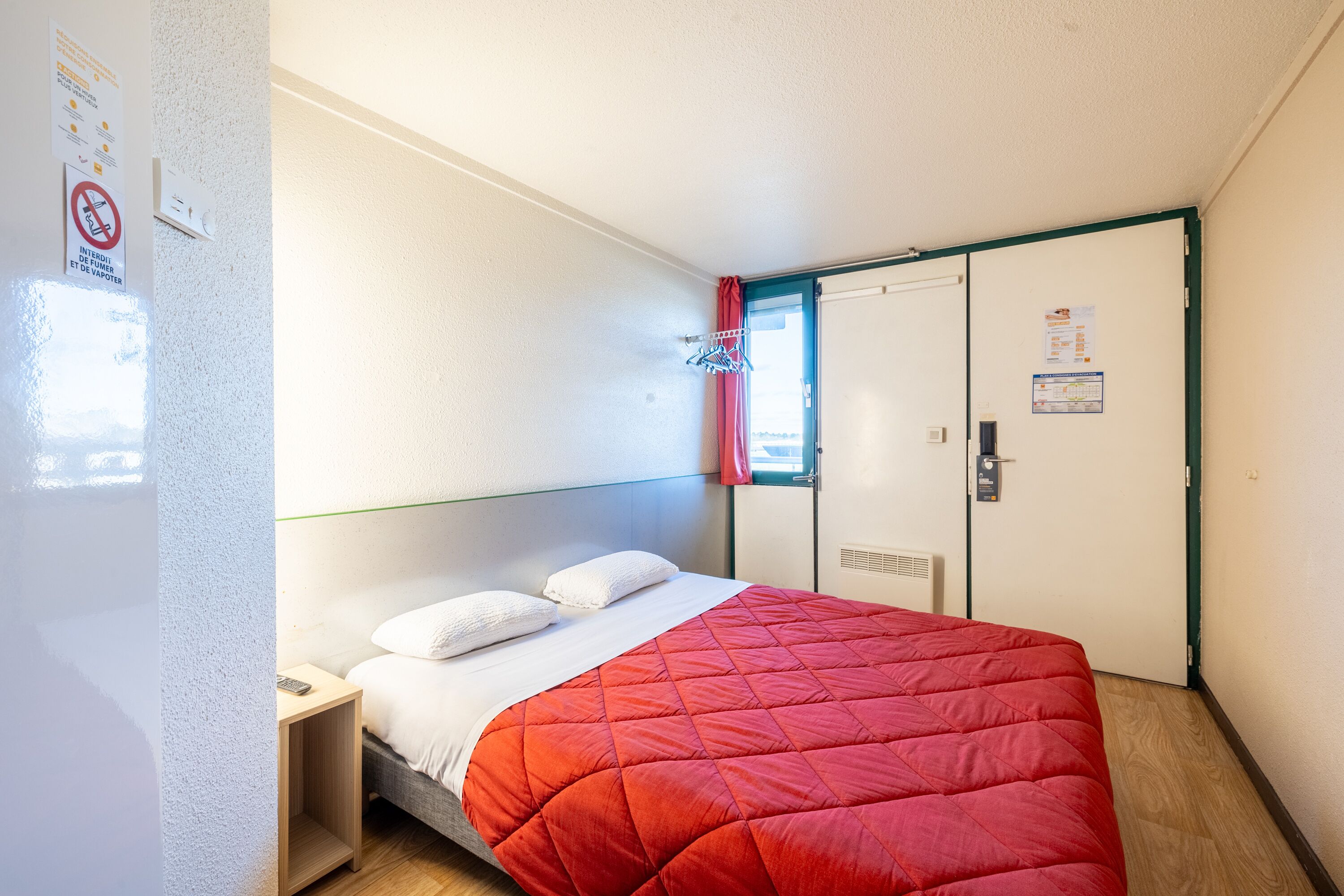 A hotel room with a bed, a nightstand, a wardrobe, and a window with a red curtain.