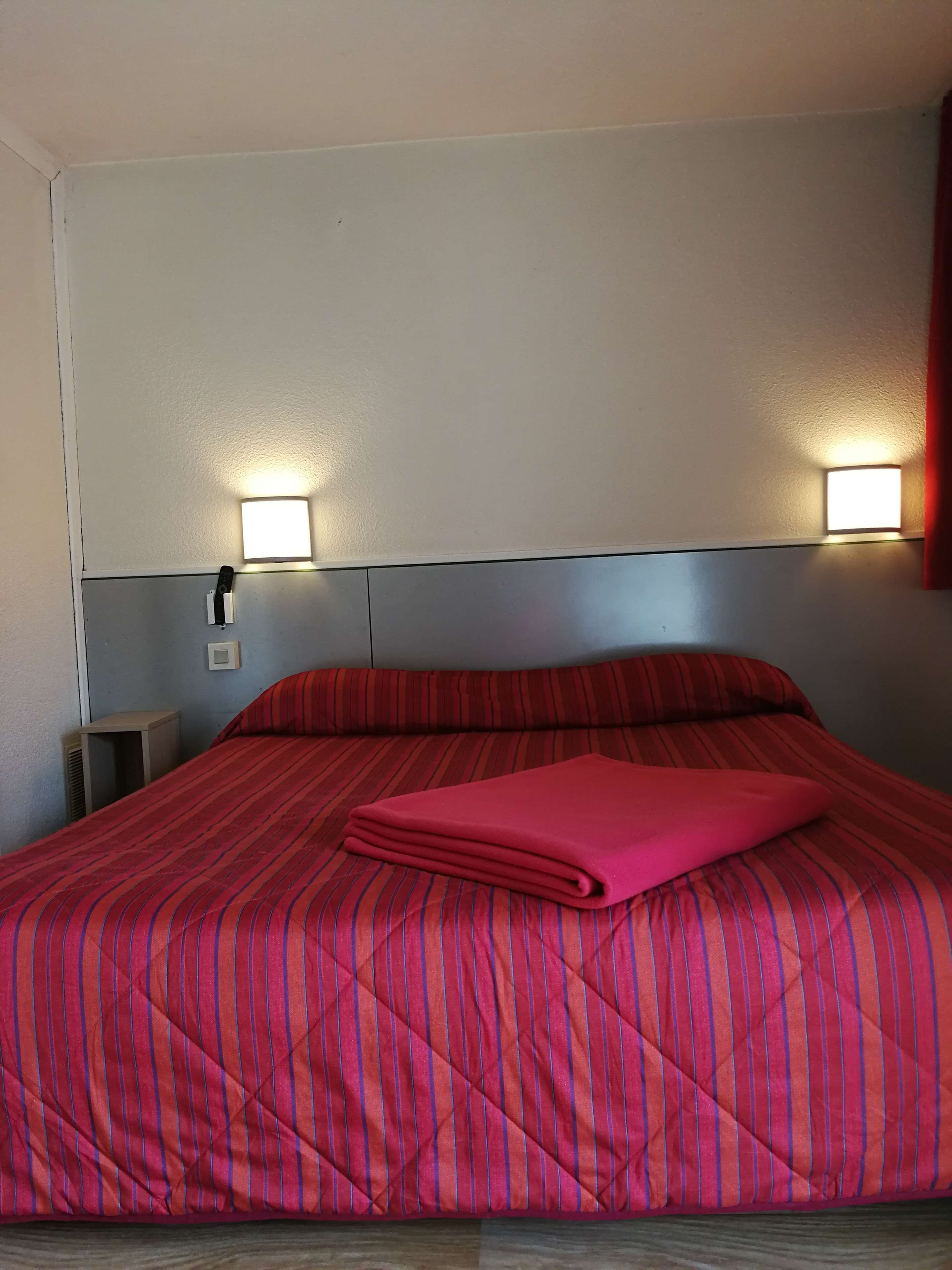 Una cama con colcha roja y azul a rayas, dos luces fijadas a la pared y una mesita de noche con lámpara.