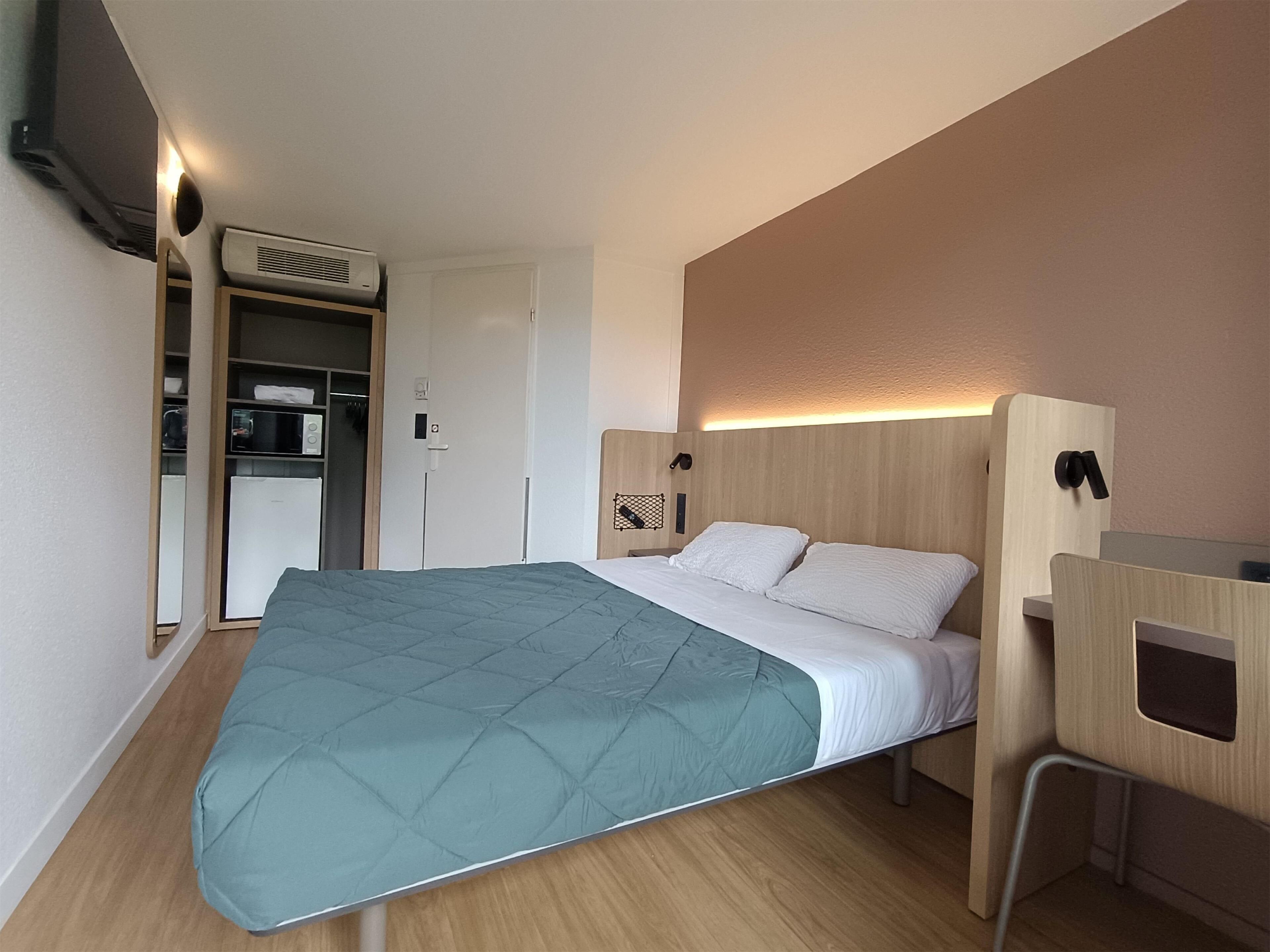 A compact hotel room with a bed, a desk, a wardrobe, and a kitchenette.