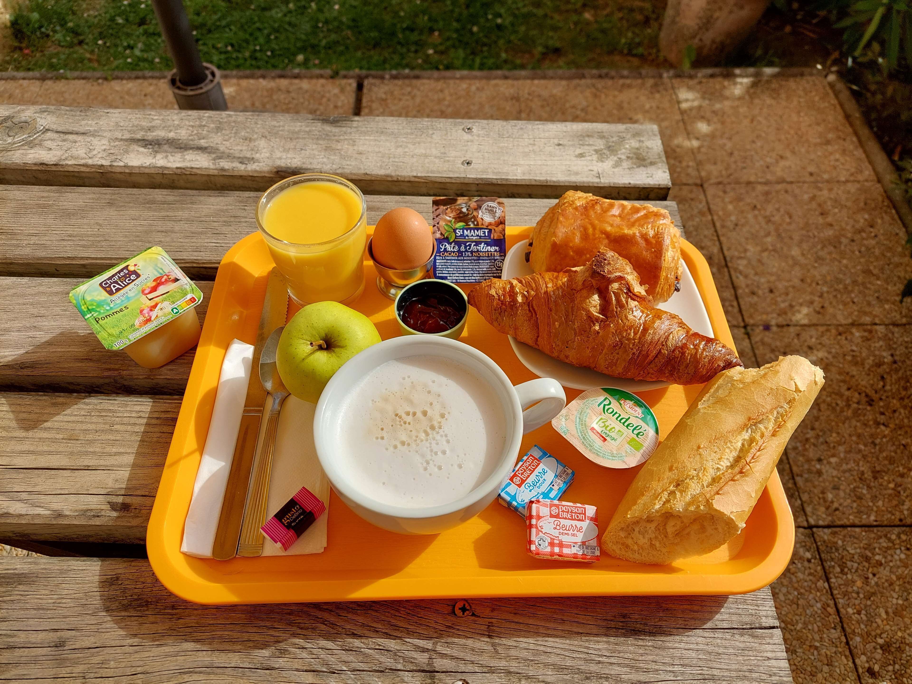 Una bandeja con alimentos para el desayuno, incluyendo un croissant, una manzana, zumo de naranja y una taza de café.