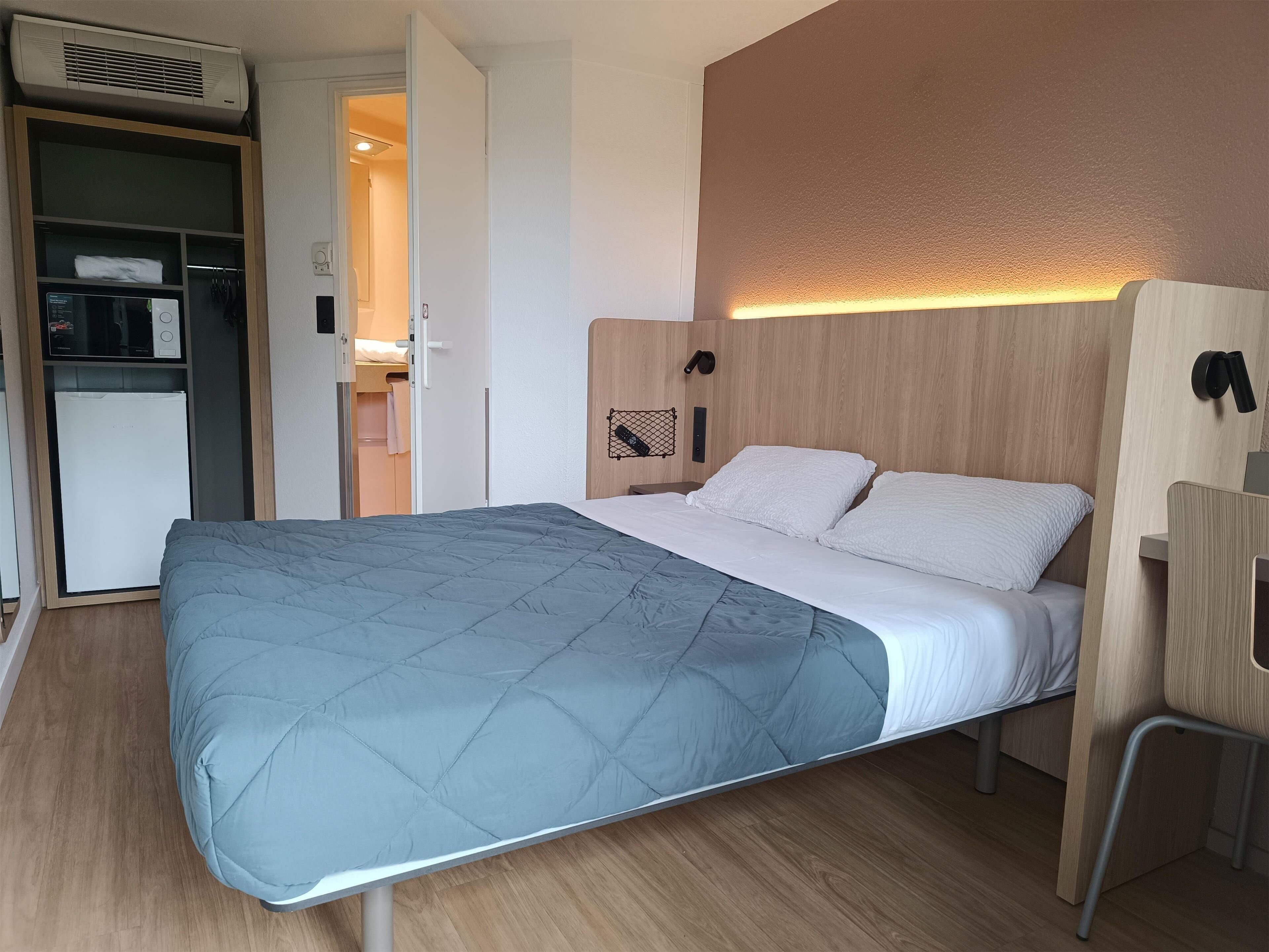 A single bed with a blue quilt, a wooden headboard, and a bedside table with a lamp.
