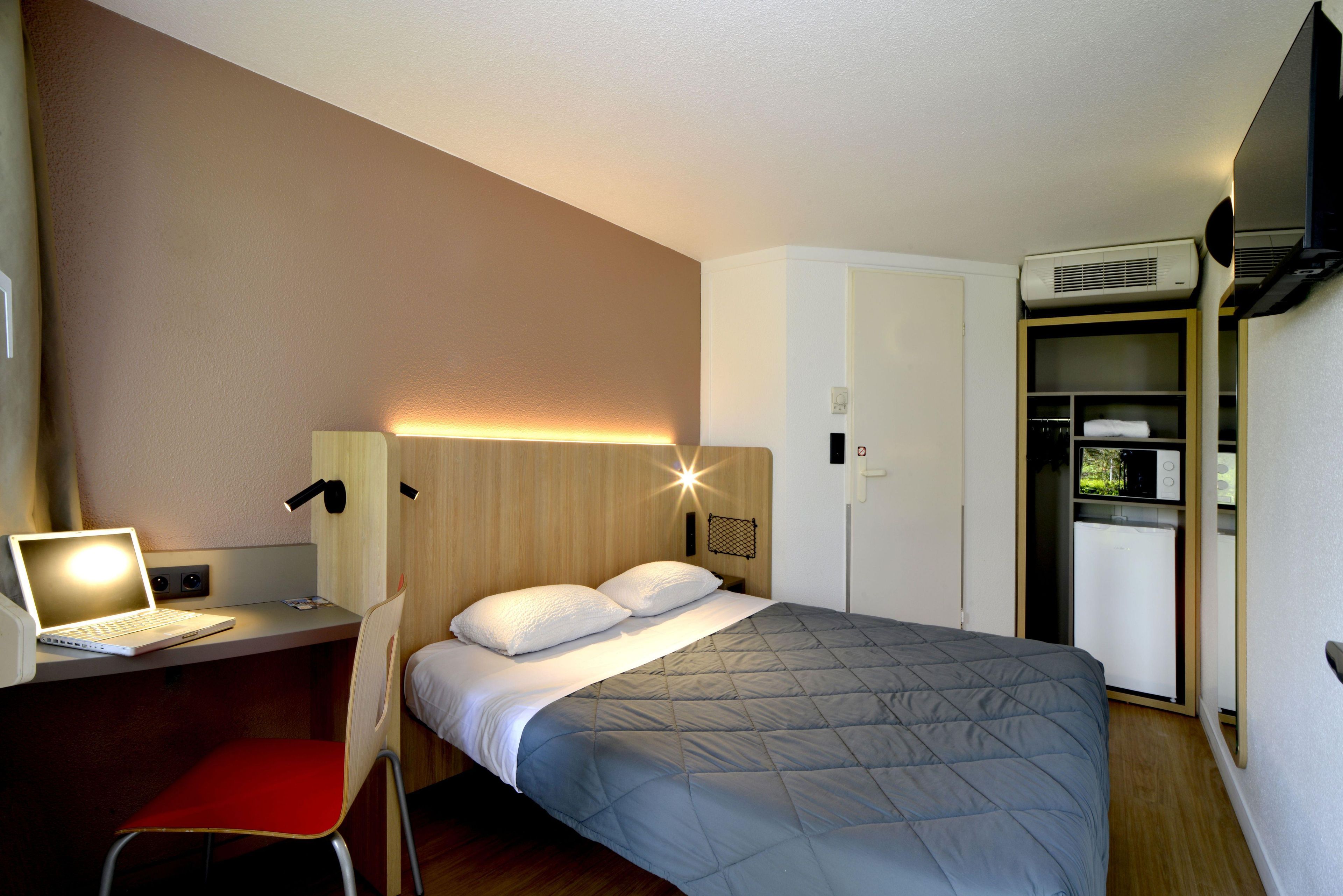 A hotel room with a bed, a desk with a laptop, a chair, a wardrobe, and an air conditioning unit.