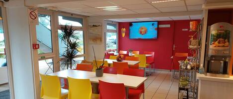 A brightly lit dining area with red and yellow chairs, a large window, and a television screen on the wall.