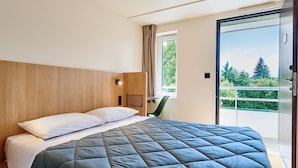 A modern hotel room with a large bed, a wooden headboard, a small desk, and a sliding glass door to a balcony.