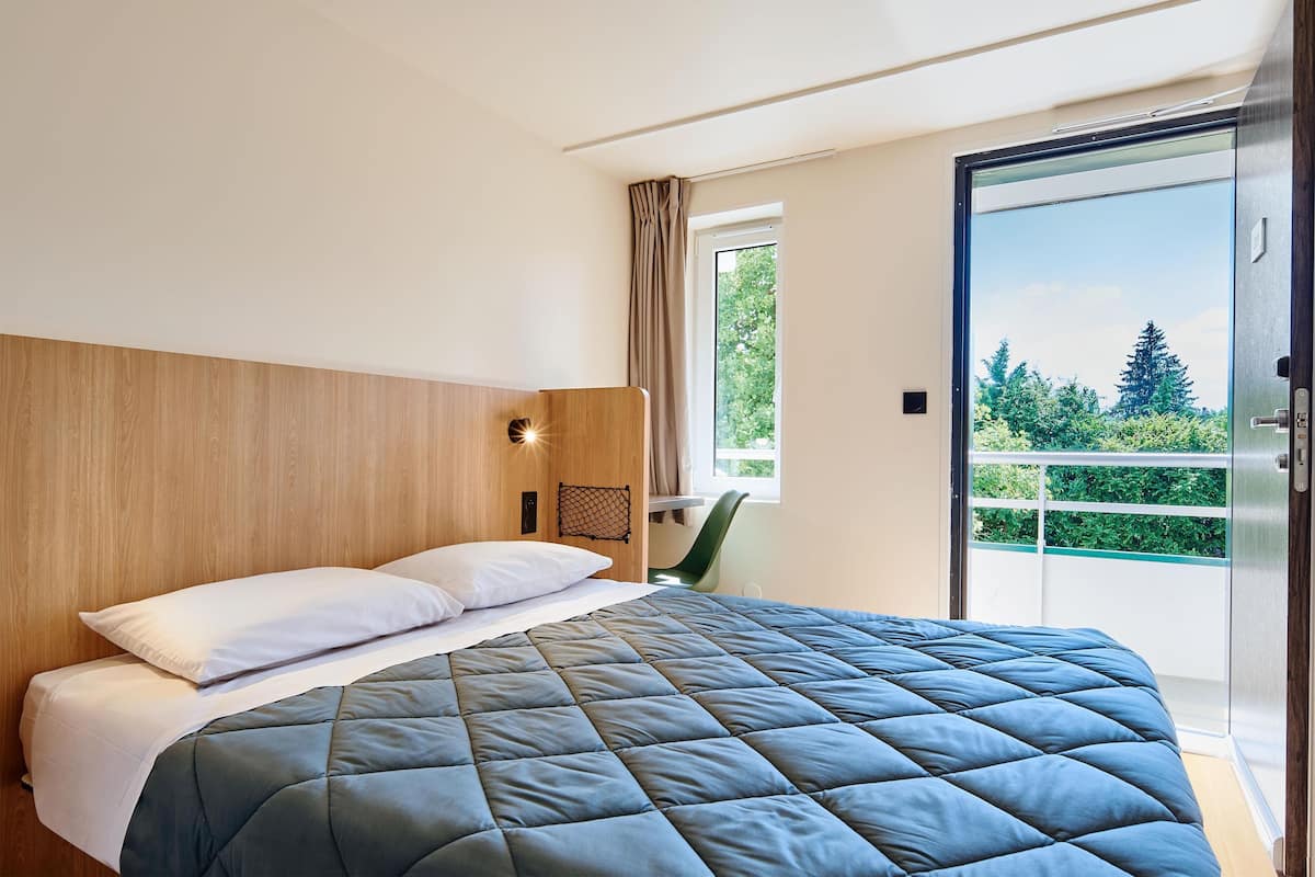 A modern hotel room with a large bed, a wooden headboard, a small desk, and a sliding glass door to a balcony.