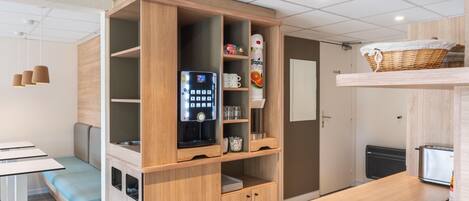 Une cuisine moderne et compacte, équipée d’une machine à café intégrée, d’un petit réfrigérateur et d’une étagère contenant les ustensiles de cuisine essentiels.