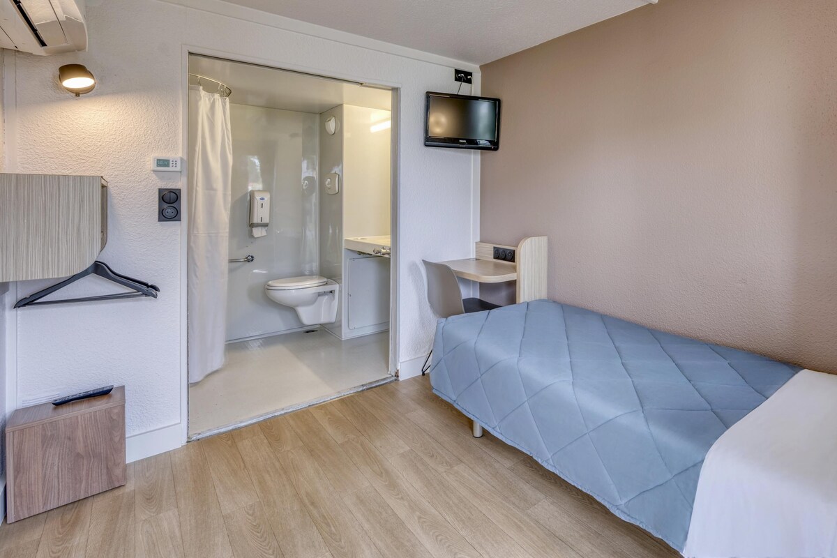 Habitación pequeña con cama, un escritorio, televisor y baño visible a través de una puerta abierta.