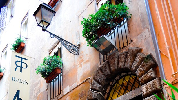A building with a stone staircase leading to a yellow door, potted plants, and a sign with a symbol and text. - Relais Palazzo Taverna (Rome)
