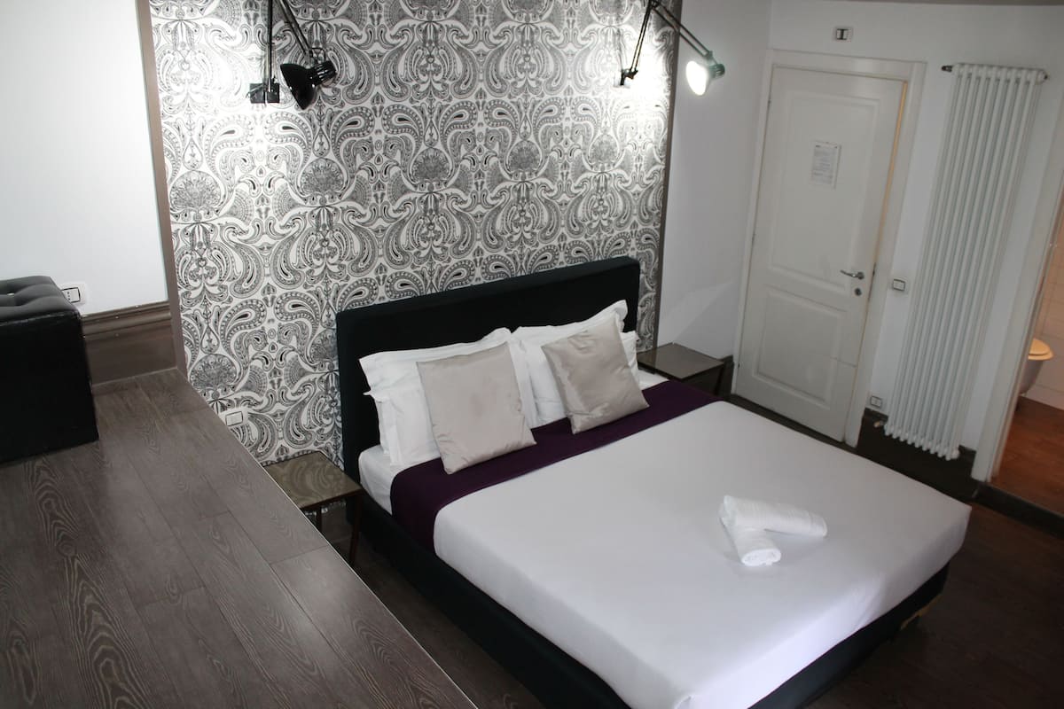 A neatly made bed with white linens and a purple blanket, a patterned wallpaper, a black headboard, and a white door with a radiator.