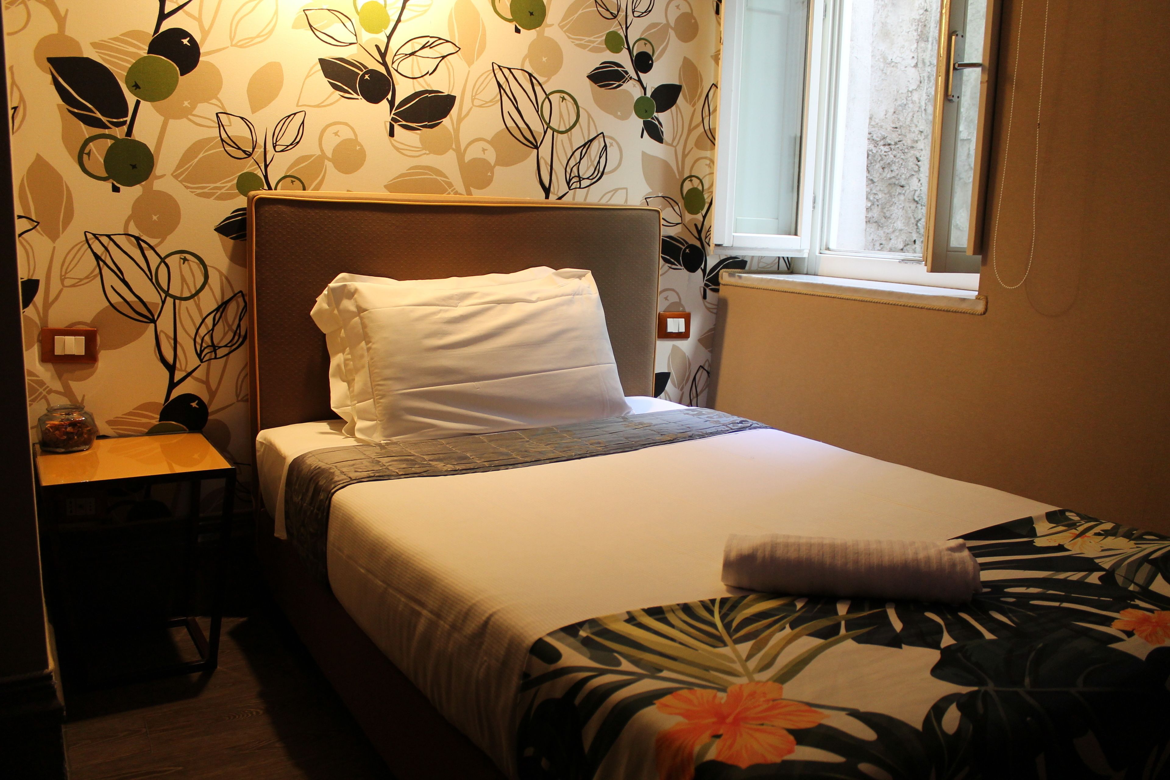 Un lit bien fait, avec un oreiller blanc et une couverture à motifs floraux, une table de chevet en bois avec une lampe, et un mur orné d’un motif de feuilles.