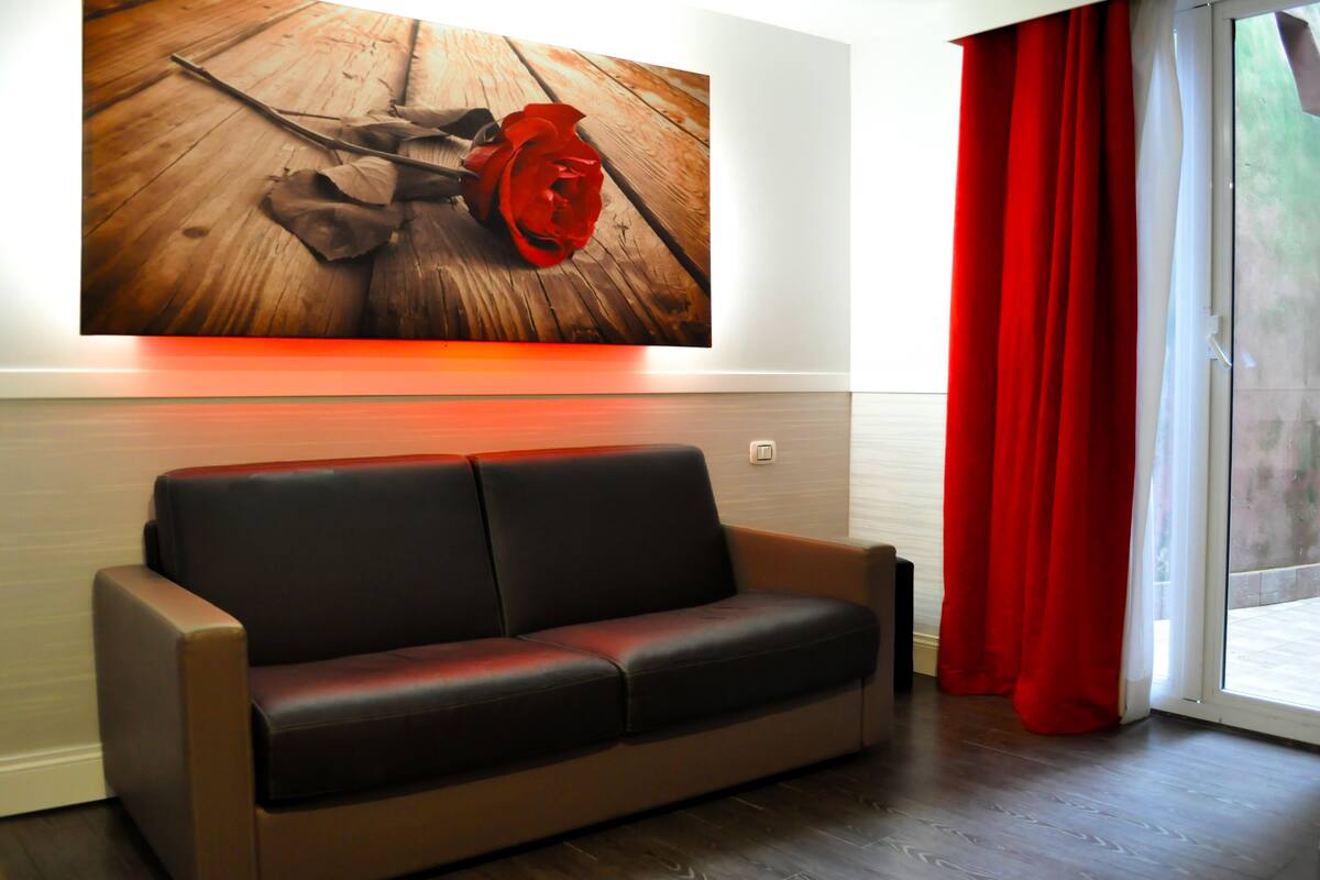 A modern living room with a black leather sofa, a wooden floor, and a large painting of a rose on the wall.