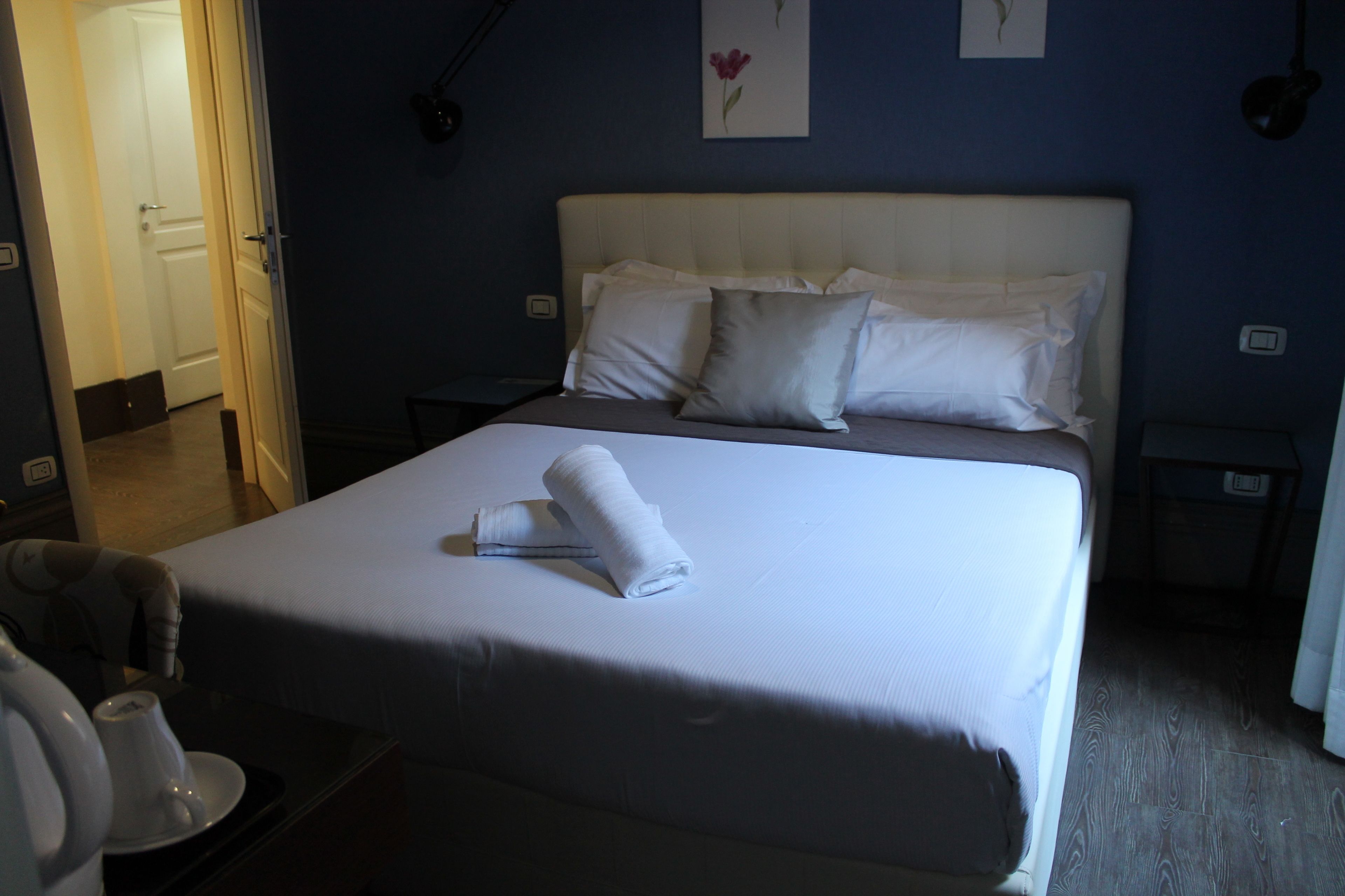 Una cama bien tendida con almohadas y una toalla enrollada, una mesita de noche con una lámpara, y una puerta que da a otra habitación.