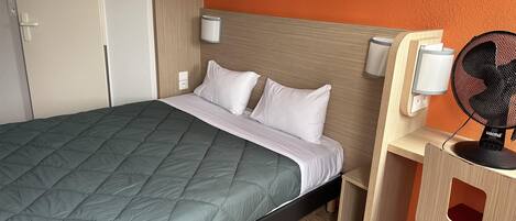 A neatly made bed with a green quilt, a wooden headboard, and a desk with a fan and lamp.