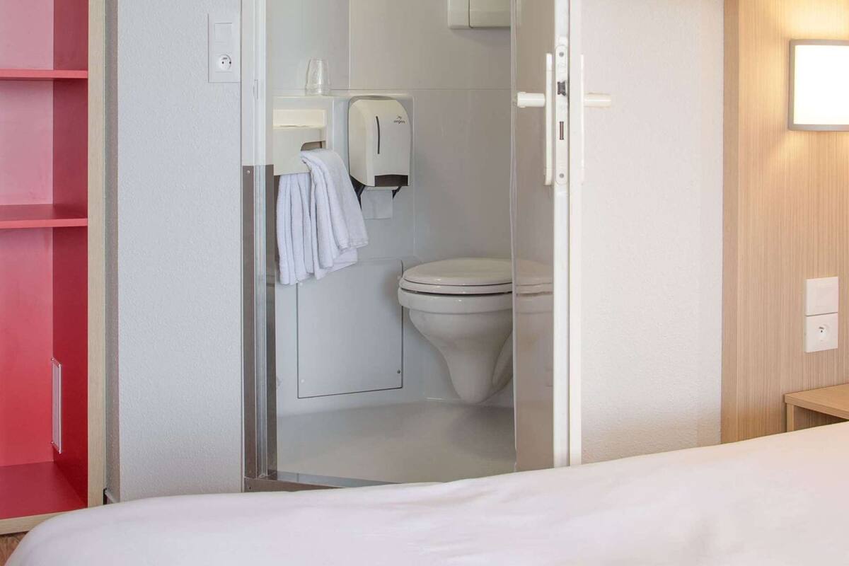 Une chambre d’hôtel avec un lit superposé, une salle de bain avec des toilettes et un sèche-mains, et un meuble de rangement rouge.