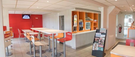 A modern cafe with a red accent wall, a TV mounted on the wall, and a counter with magazines and a coffee machine.