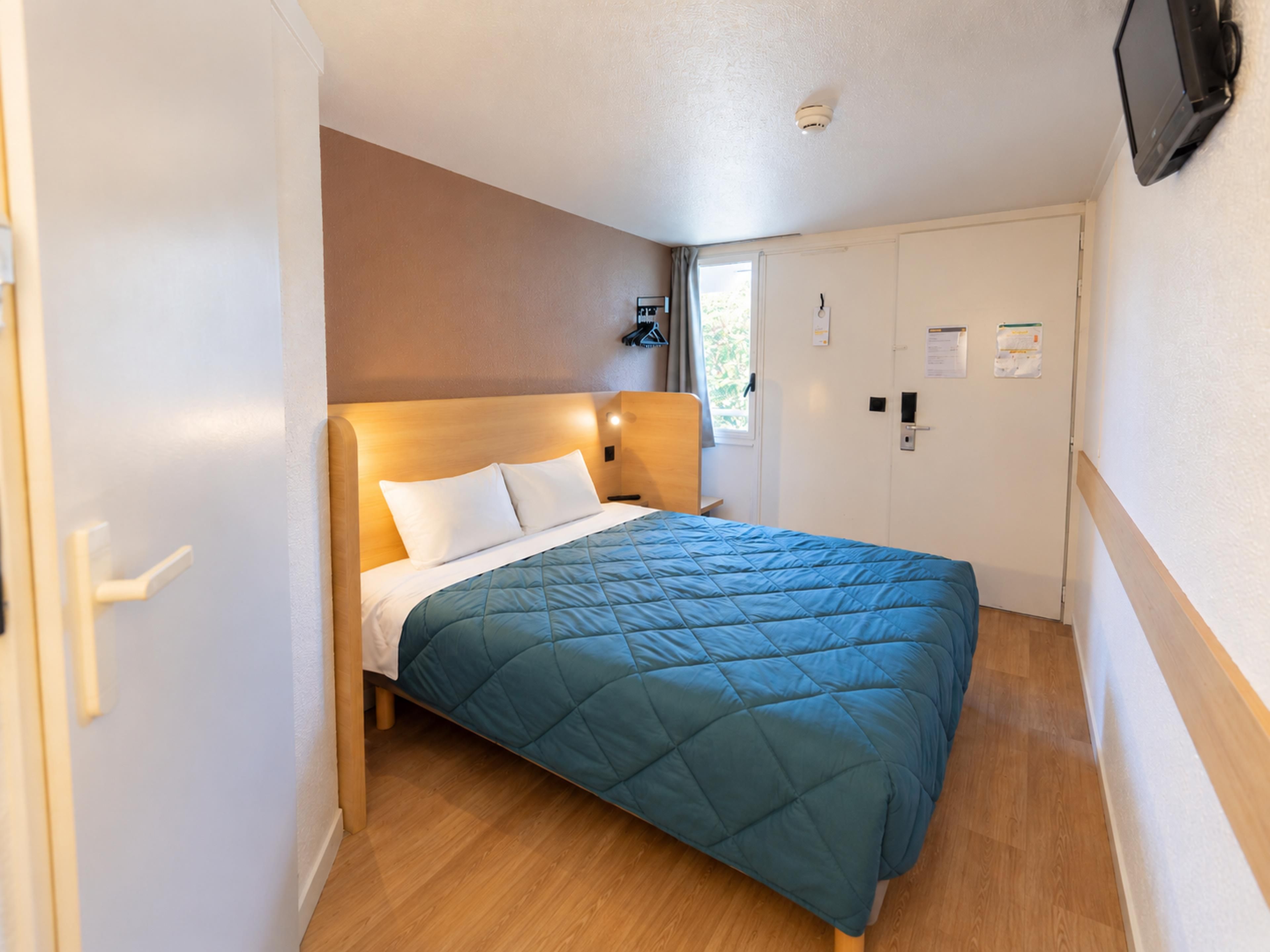 Une chambre d’hôtel avec un lit, un plancher en bois, une porte blanche, un téléviseur fixé au mur et une fenêtre avec des rideaux.