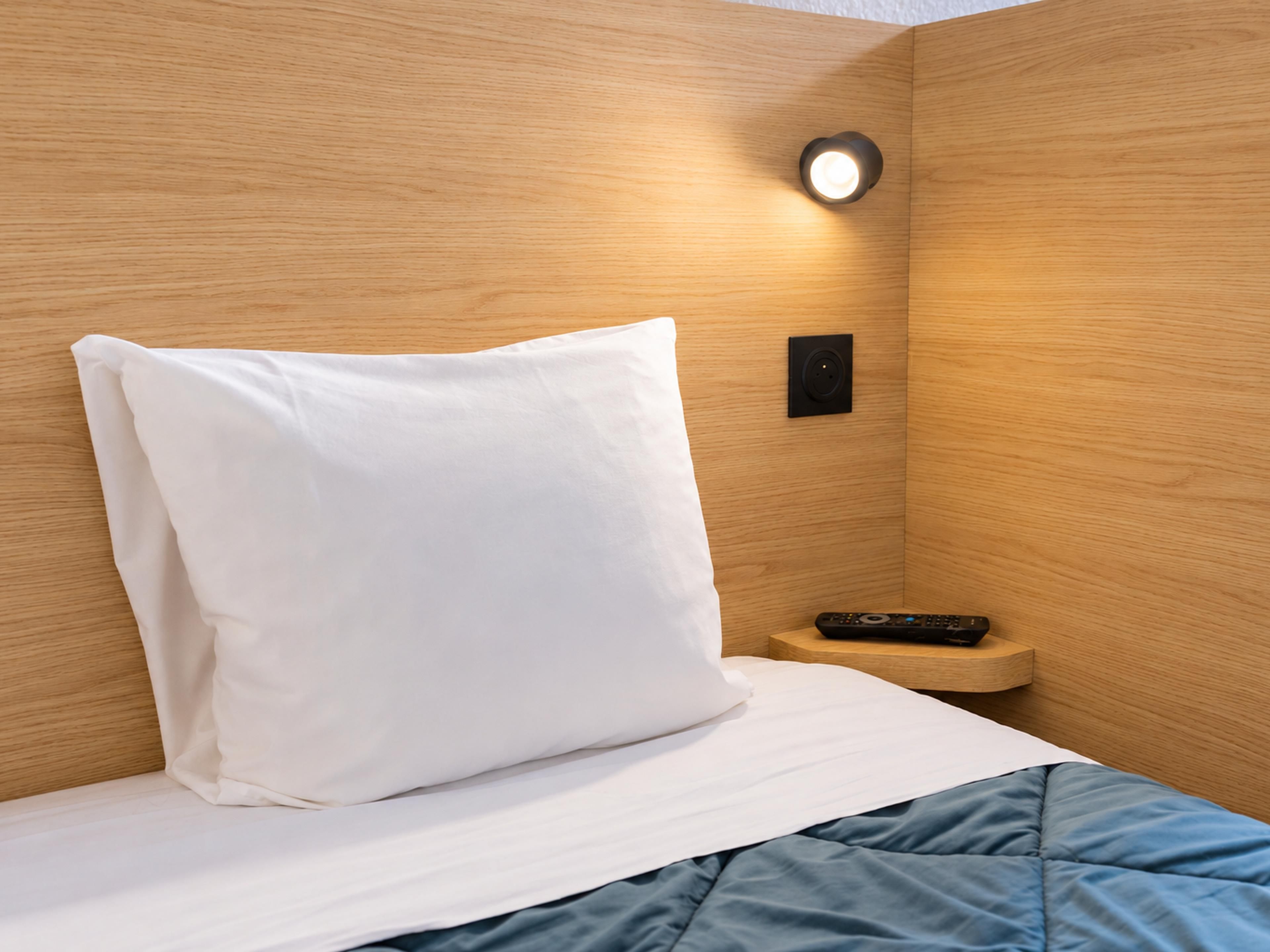 Una cama bien hecha con un almohadón blanco y una manta azul, una mesita de noche con un control remoto y una lámpara fijada a la pared.