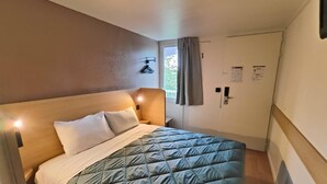 A single bed with a wooden headboard, a quilted green bedspread, and two pillows. There is a nightstand with a lamp, a window with curtains, and a door with a lock. - Premiere Classe Tarbes - Bastillac (Tarbes)