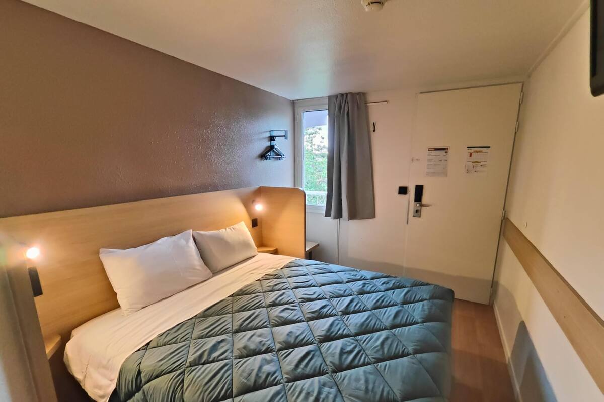 Een eenpersoonsbed met houten hoofdbord, een groene gewatteerde spreidek en twee kussens. Er is een nachtkastje met een lamp, een raam met gordijnen en een deur met slot.