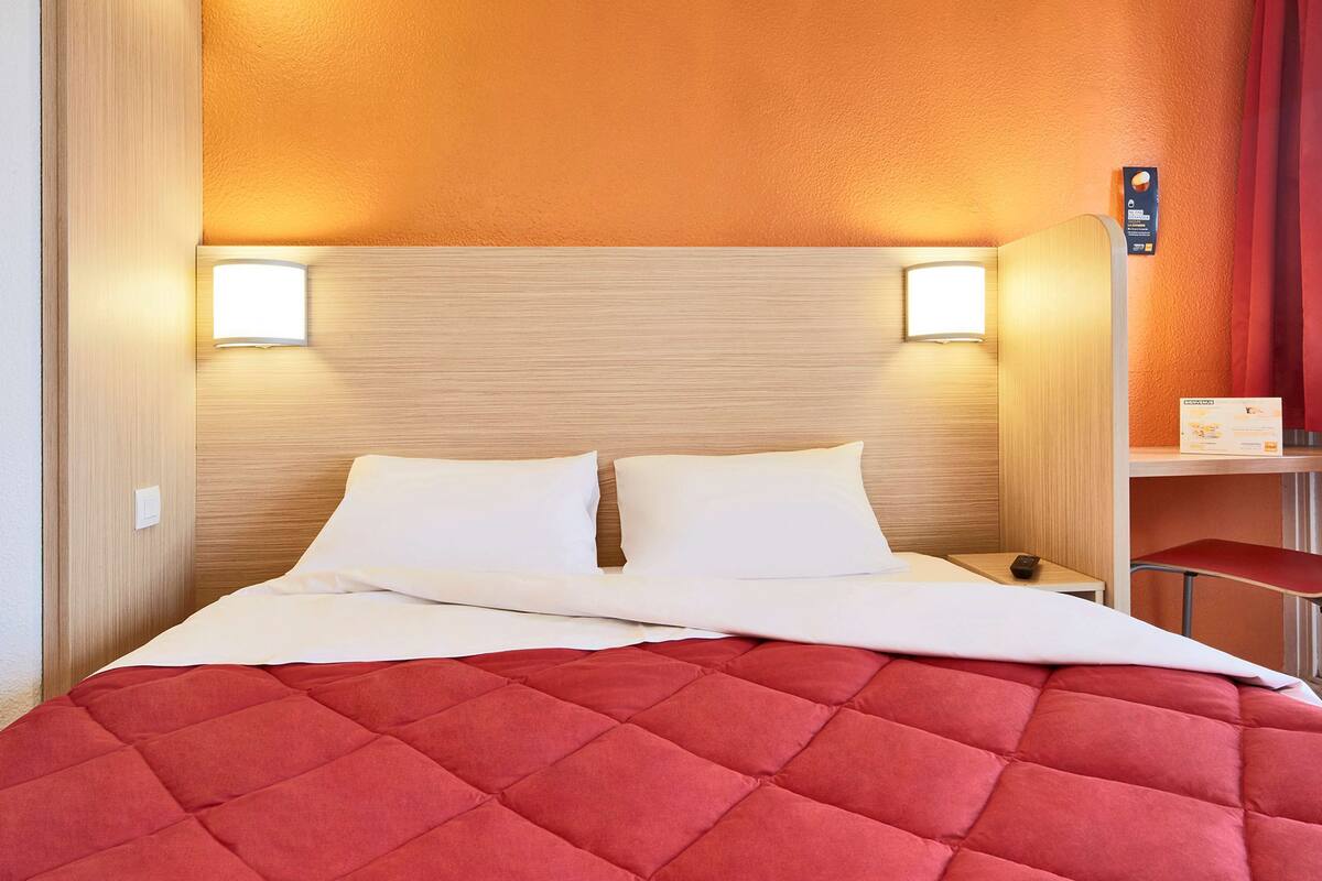 Ein Hotelzimmer mit einem Bett, zwei Nachttischlampen, einem roten, gepolsterten Kopfteil und einem kleinen Tisch mit einer Fernbedienung.