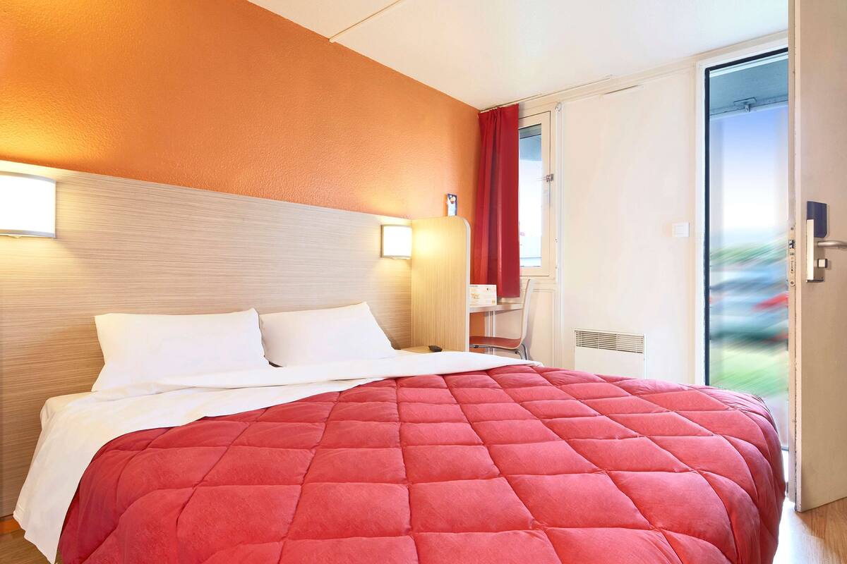 Ein Hotelzimmer mit Bett, Nachttisch, Lampe und Blick ins Freie.