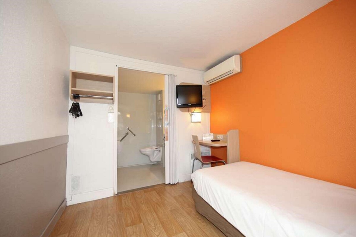 Quarto de hotel com cama, escrivaninha, televisão e zona de duche.
