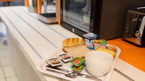 Bandeja con alimentos para el desayuno, incluyendo pan, yogur y mermeladas, sobre una encimera cerca de una cafetera.