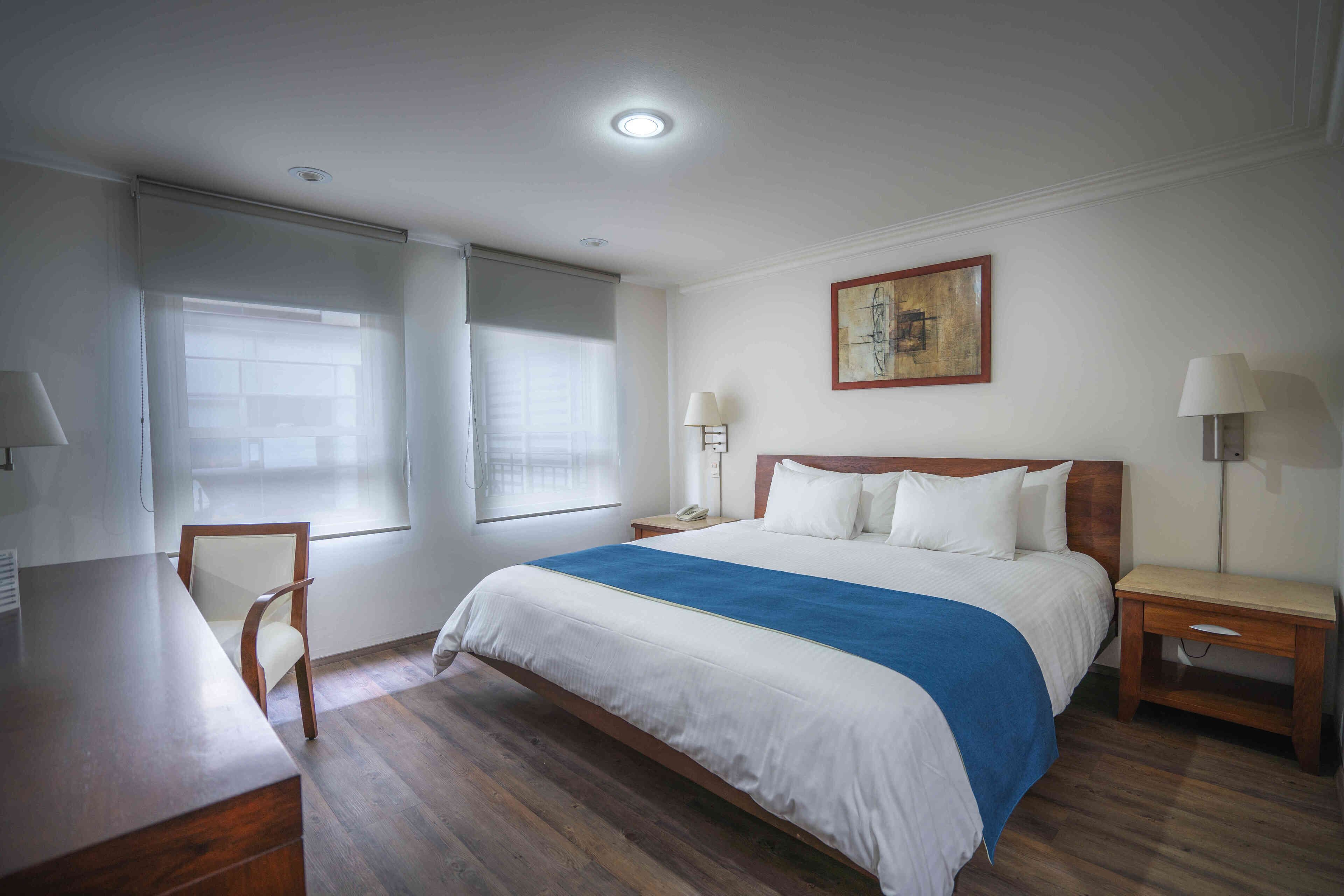 A hotel room with a bed, bedside tables, a desk, a chair, and a window with blinds.