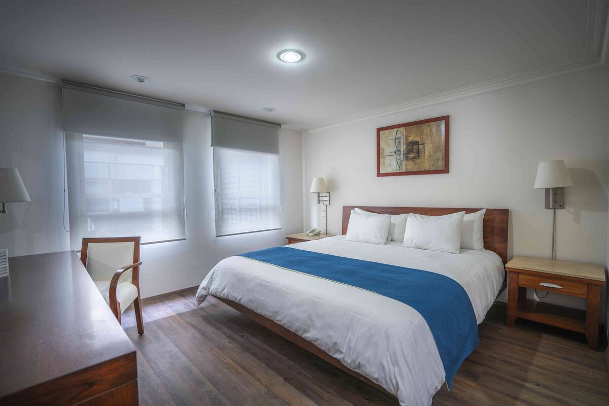 A hotel room with a bed, bedside tables, a desk, a chair, and a window with blinds.
