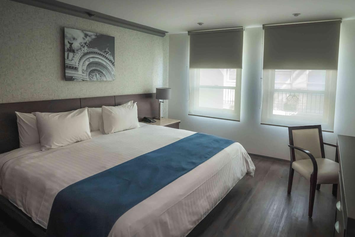 A hotel room with a large bed, a nightstand, a chair, a lamp, and a window with blinds.