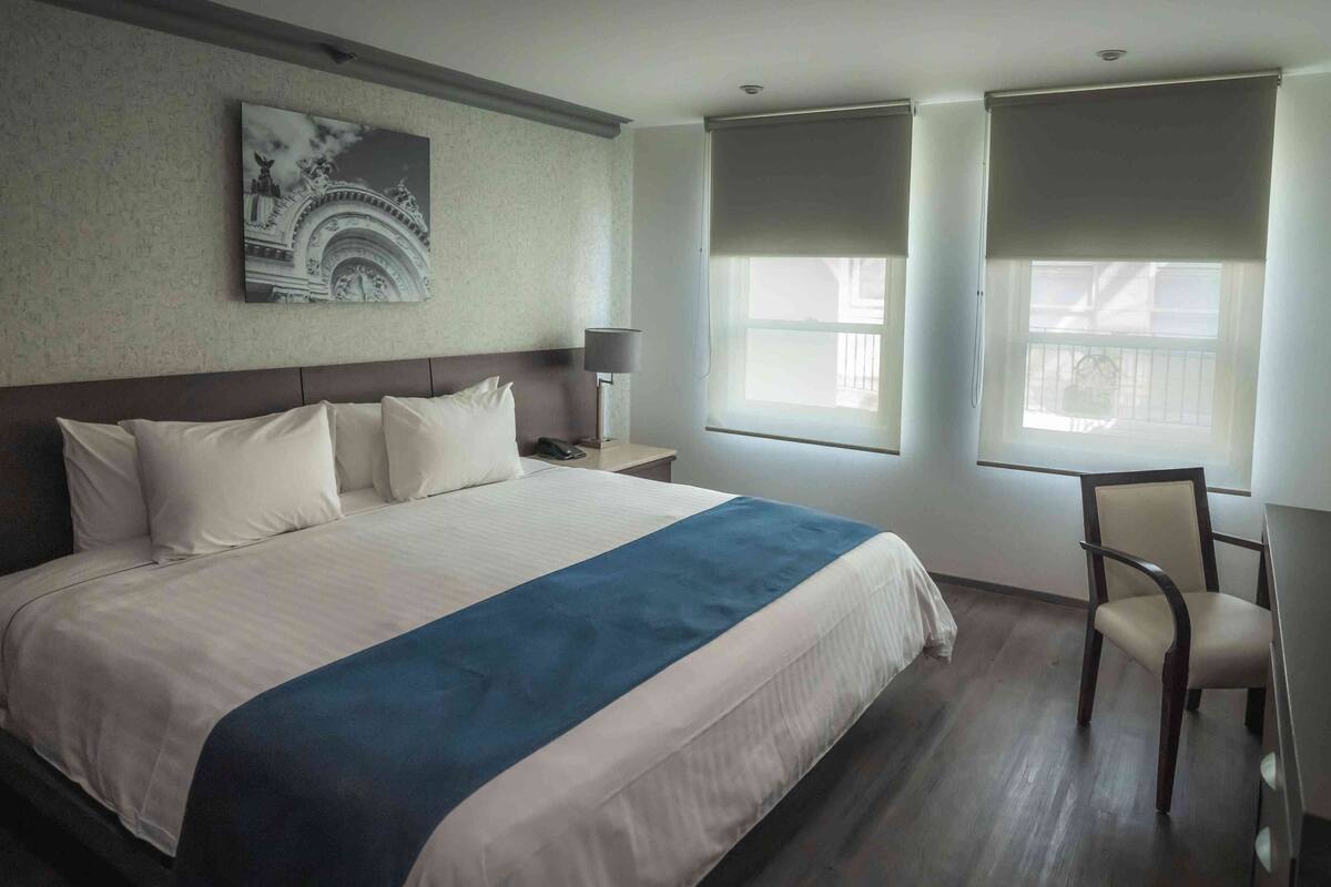 A hotel room with a large bed, a nightstand, a chair, a lamp, and a window with blinds.