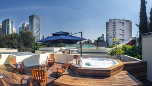 A rooftop deck with wooden furniture, a hot tub, and a view of city buildings. - Suites Obelisk (Mexico City)